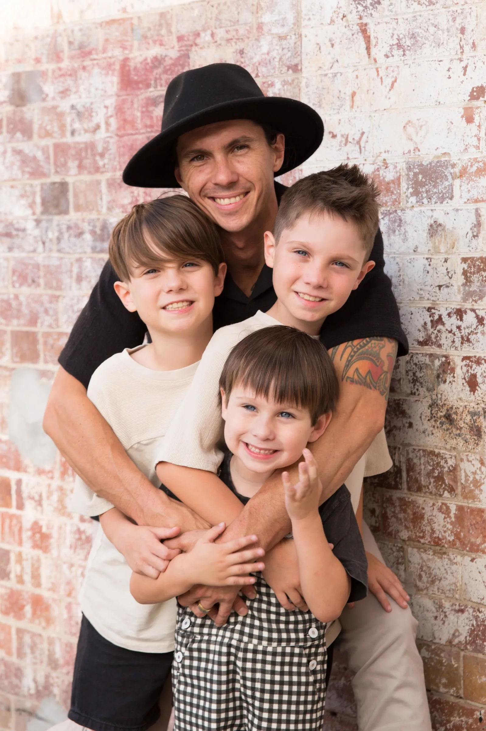 Dad in a black hat hugging his three boys by a brick wall