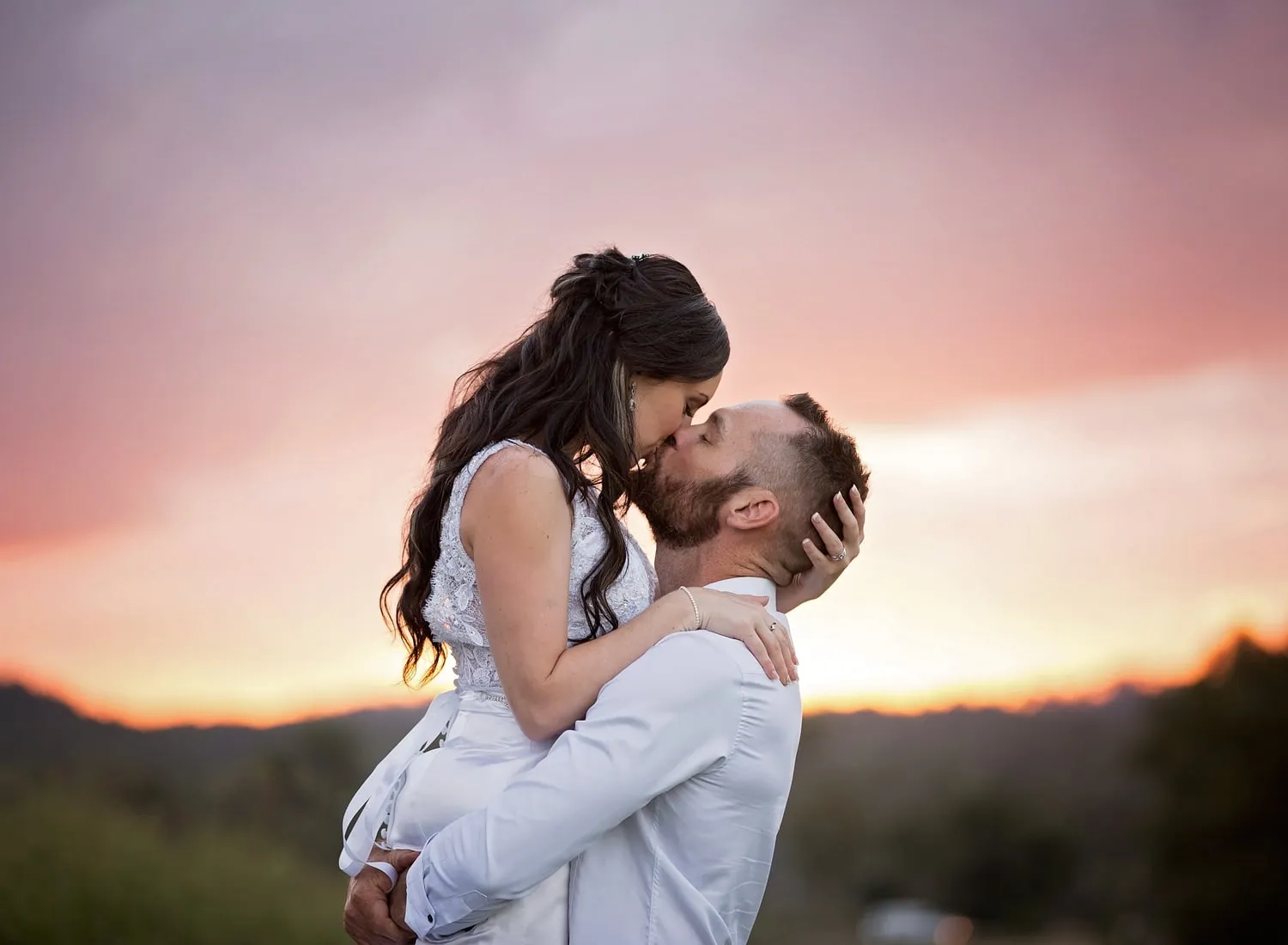 A bride and groom kissing under a pink sunset sky