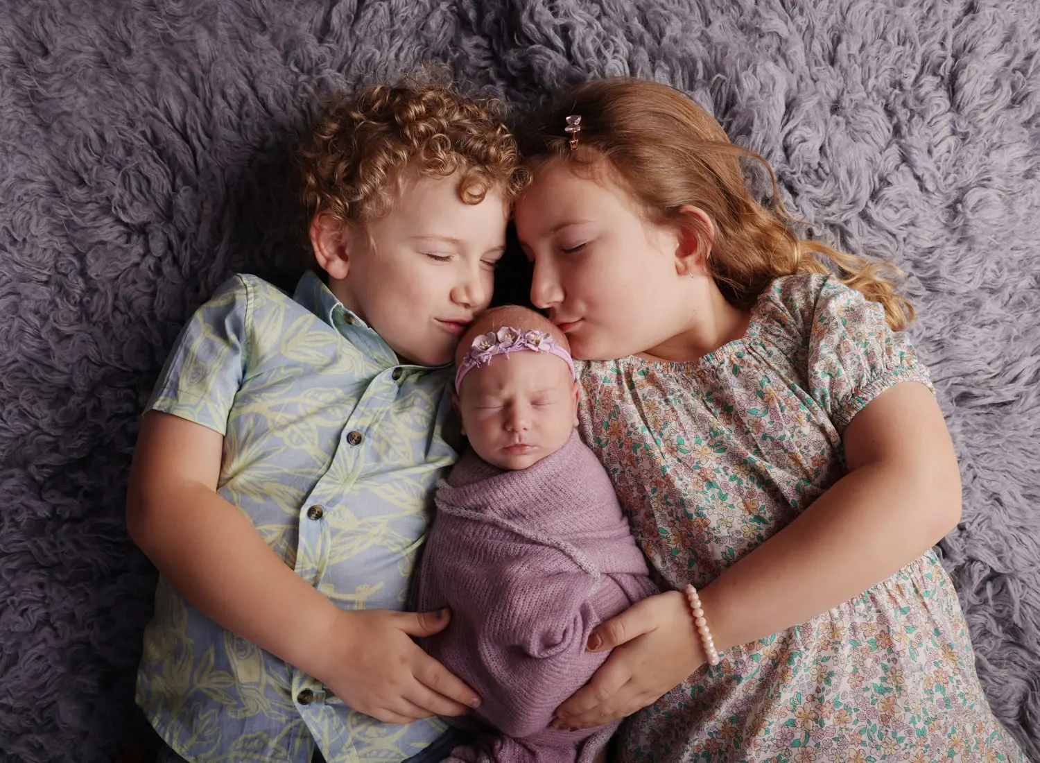 Brother and sister cuddling their newborn sister on a fluffy rug