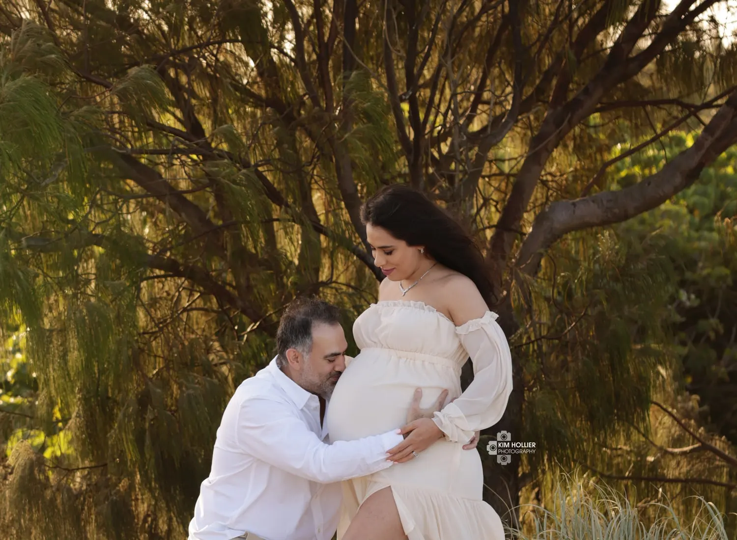 Dad kissing mum's bump under a tree at sunset